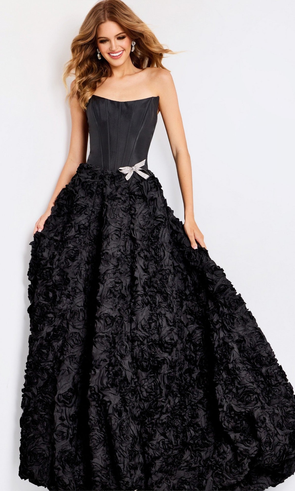 A woman models the Jovani 47695 long prom dress, showcasing its strapless corset bodice, dramatic 3D rosette skirt, and silver bow detail against a plain white background.